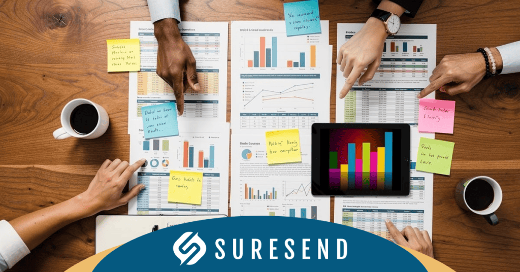 ALT: Team analyzes financial charts with coffee, tablet showing CRM data; reviewing HubSpot alternative Suresend. Suresend logo shown. Caption: Business team reviews CRM alternatives to HubSpot using financial charts and digital analytics with Suresend branding visible. Description: Four professionals collaborate on printed financial documents and an e-tablet displaying a colorful CRM bar graph, researching top alternatives to HubSpot. Coffee cups and the Suresend logo signal teamwork and highlight the Sure Send CRM Resource Center as a key resource for efficient business management solutions.