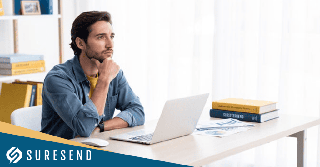 A man sits at a desk with a laptop, looking thoughtful. There are documents and books nearby as he considers optimizing his workflow with the Best Real Estate CRM with Pipeline Management. The Suresend logo is in the bottom left corner. Shelves with books and decor are in the background.